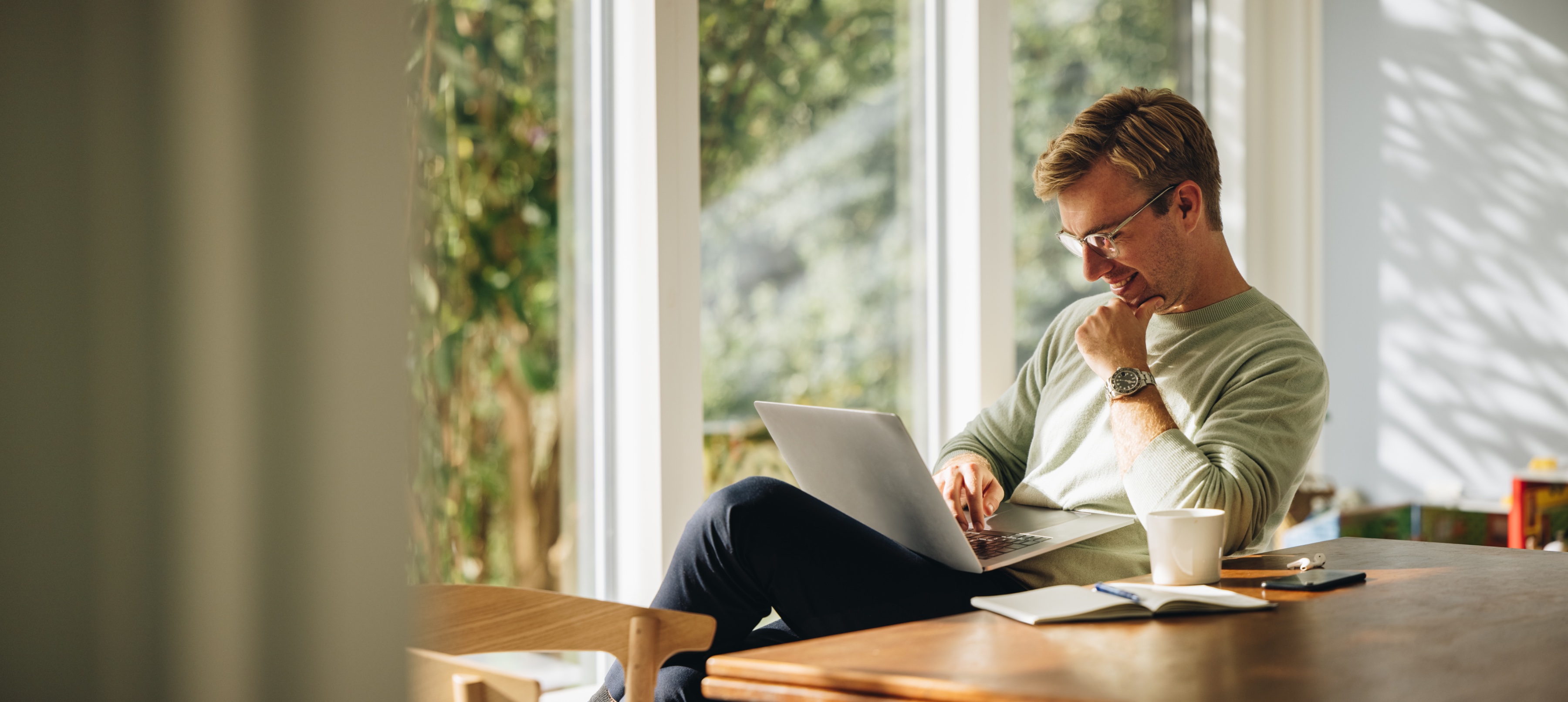 A man working on a laptop at a sunlit table by large garden windows at home
